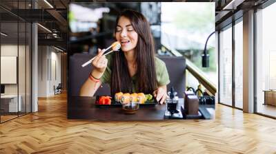 Young woman eating and enjoying fresh sushi in luxury restaurant. Female client holding food sticks and eating oriental meal on lunch. Concept of food. Wall mural